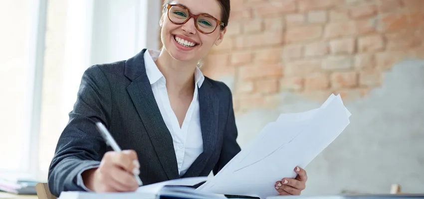 Smiling woman in a blazer reviewing and signing paperwork at a desk