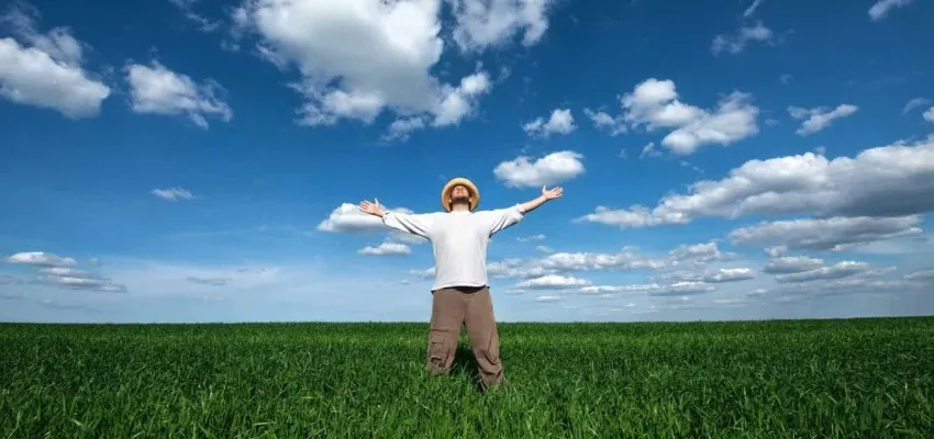 Person standing with arms outstretched in a green field under a blue sky