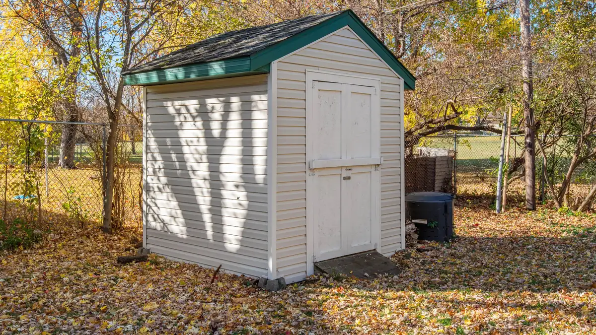 Small white backyard shed with double doors and a green trim roof among autumn trees