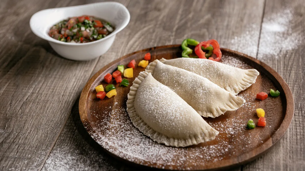 Empanadas dusted with powdered sugar on a wooden plate beside a small bowl of salsa