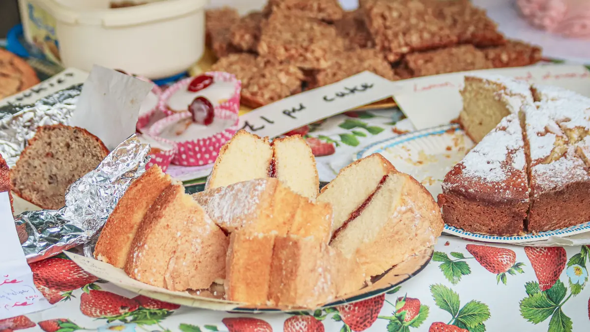 Homemade sponge cake slices and other baked goods on a market stall table with a strawberry-print cloth