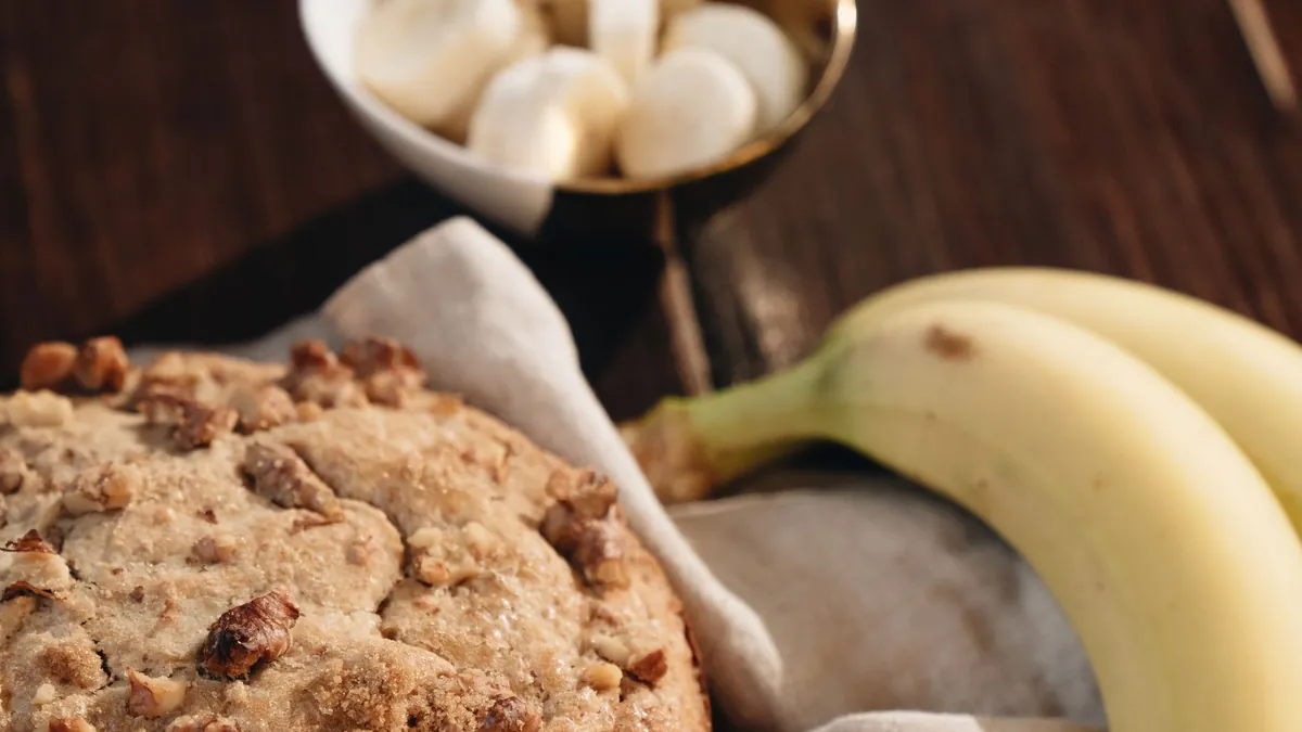 Round homemade banana bread with walnuts beside fresh bananas on a wooden table