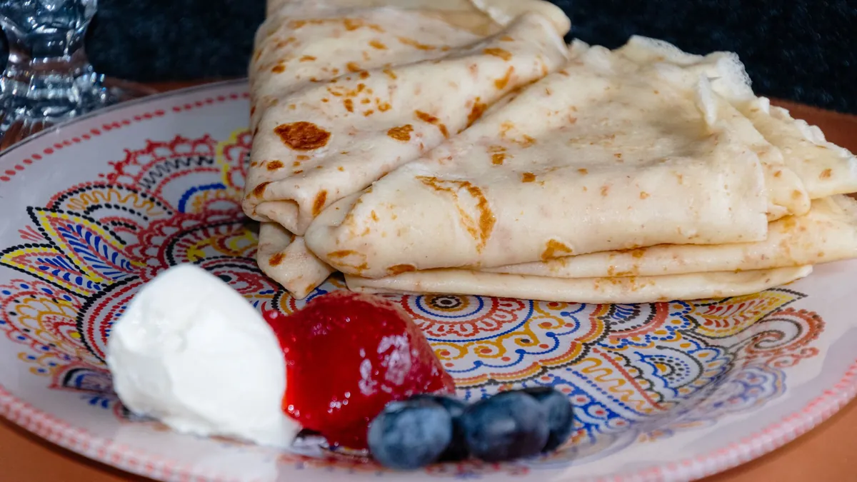 Folded crepes on a patterned plate with cream, berry sauce and blueberries