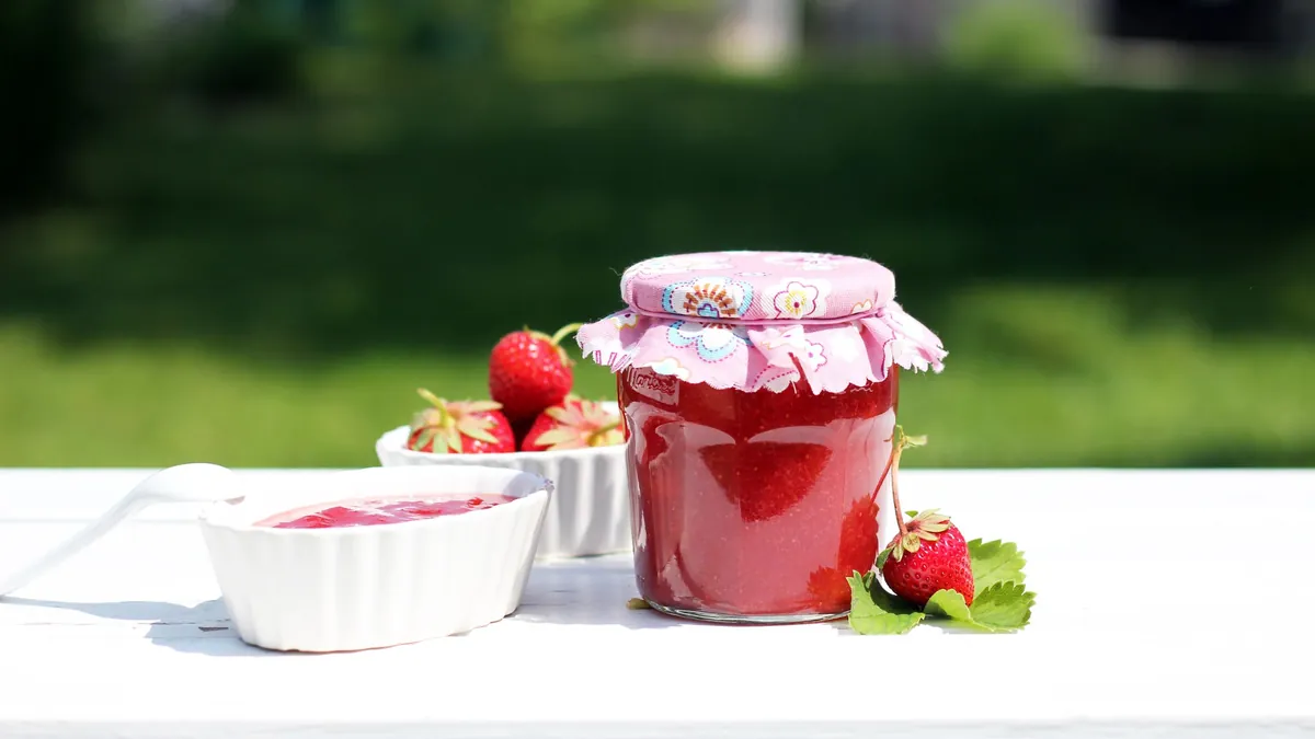 Jar of homemade strawberry jam with a fabric lid and fresh strawberries on a white table