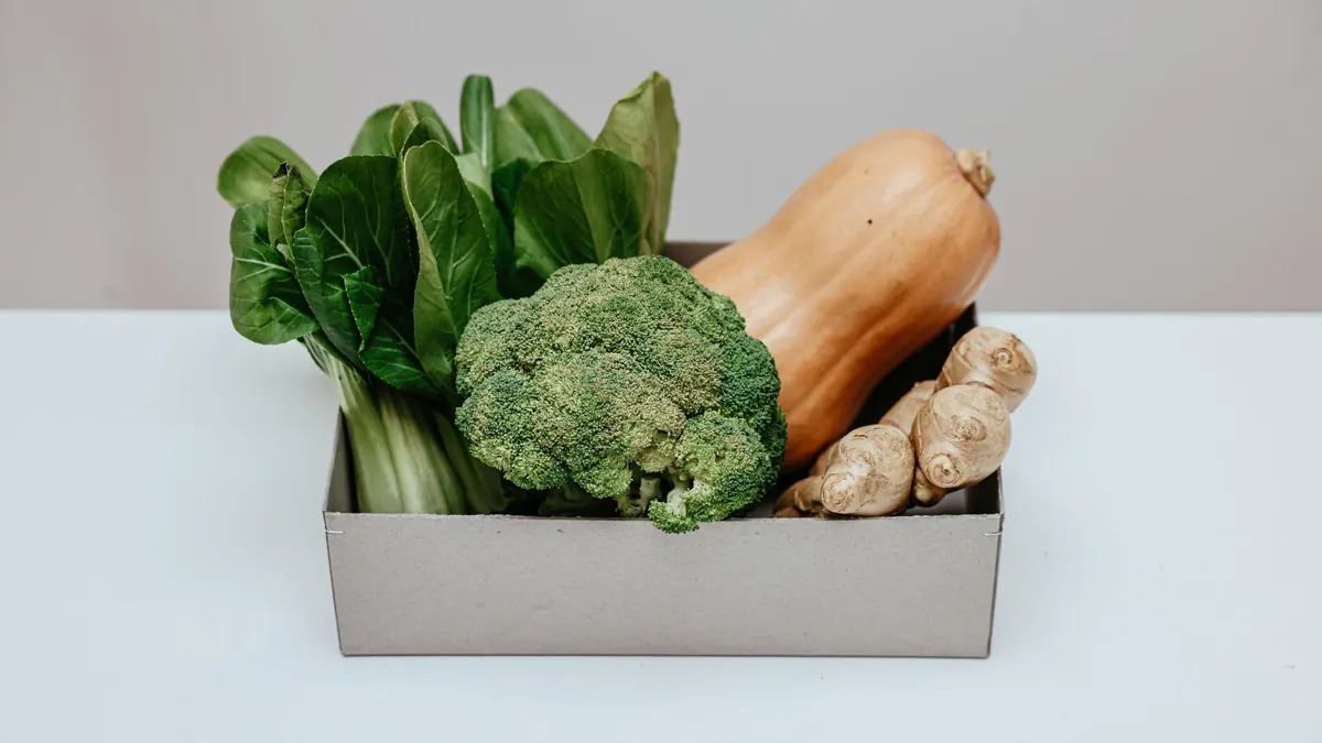 Cardboard box holding broccoli, leafy greens, a butternut squash and ginger on a white table