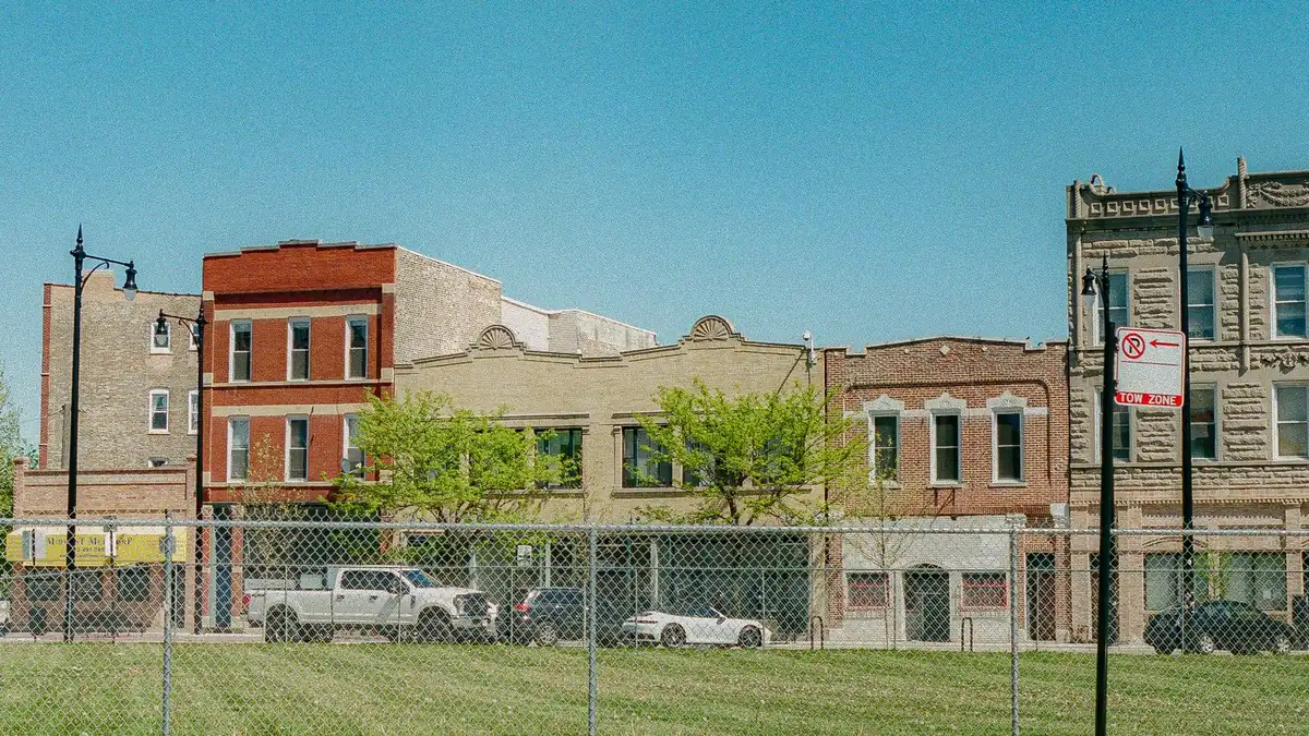 Grassy vacant lot behind a chain-link fence beside brick buildings on a city street