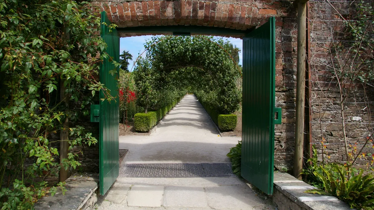Open green garden gate in a brick wall leading to a hedge-lined path