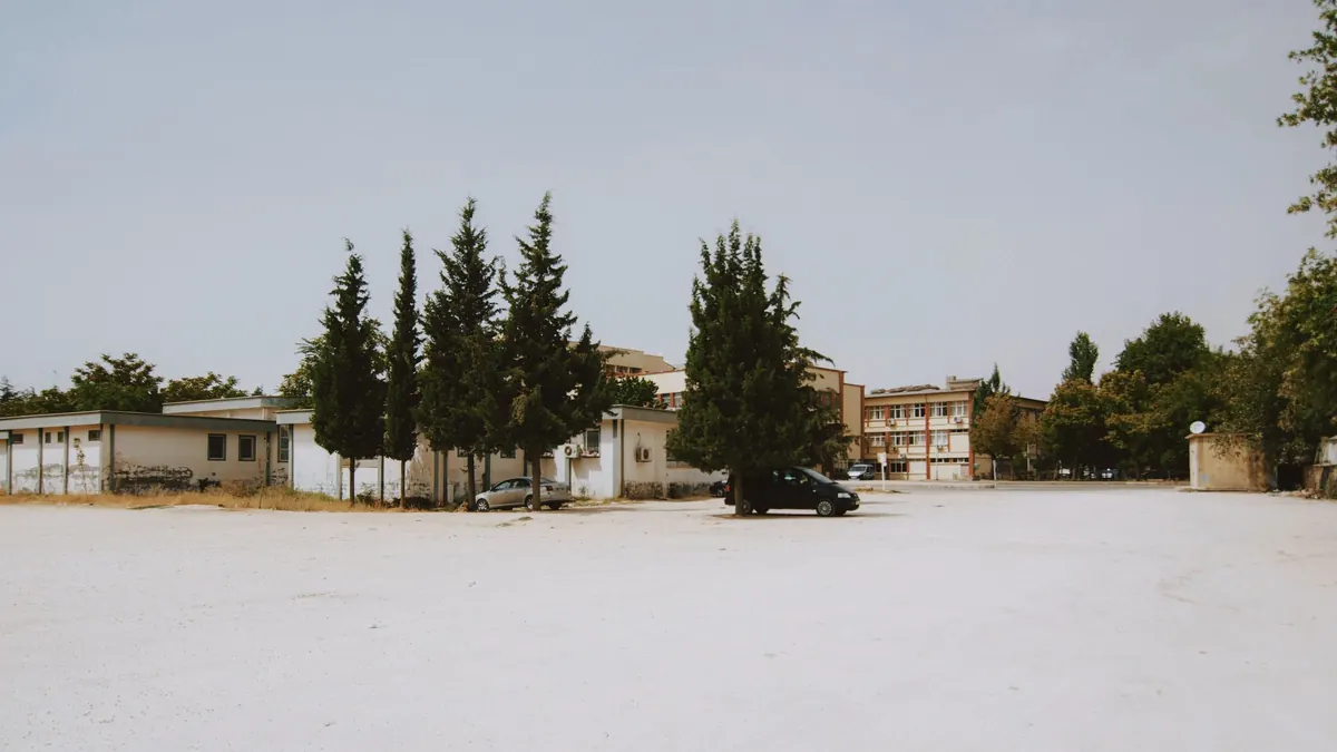 An empty paved urban lot with trees and low buildings behind it